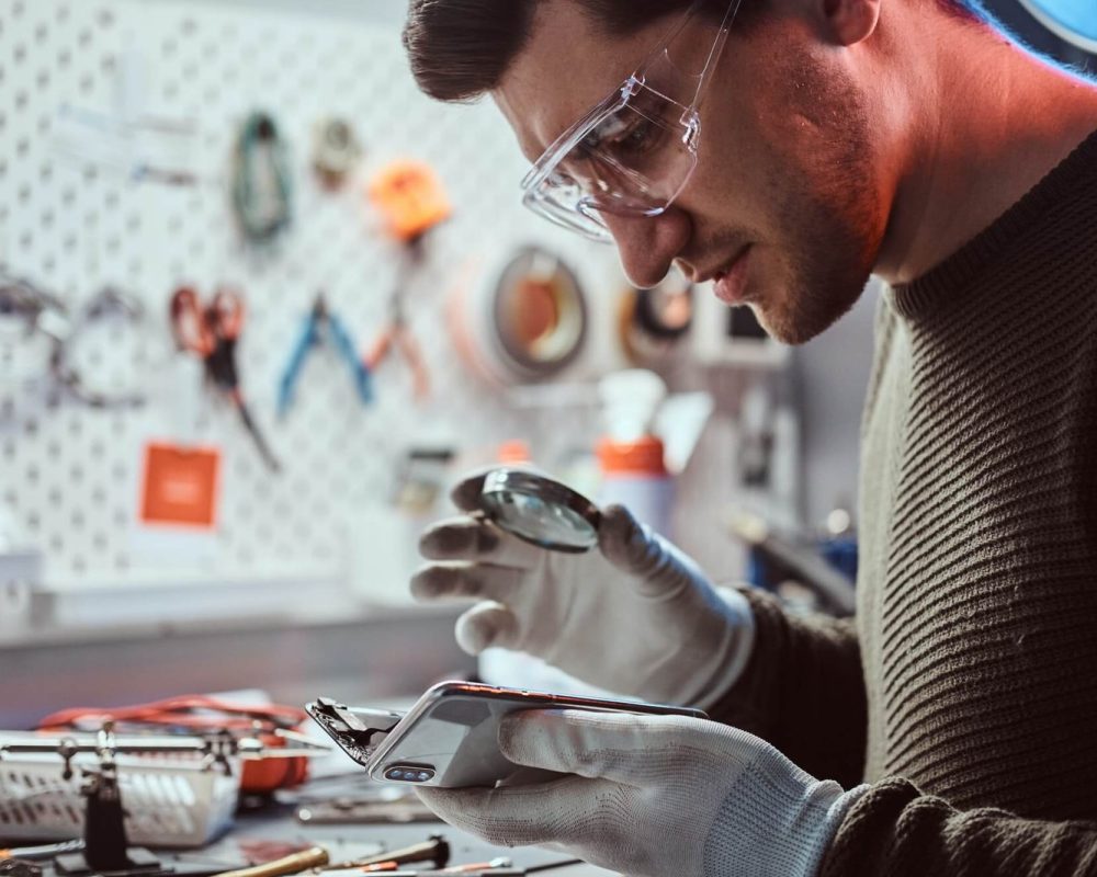 Man wearing gloves and safety glasses examines a smartphone with a magnifying glass in a workshop, surrounded by tools and equipment, focused expression.