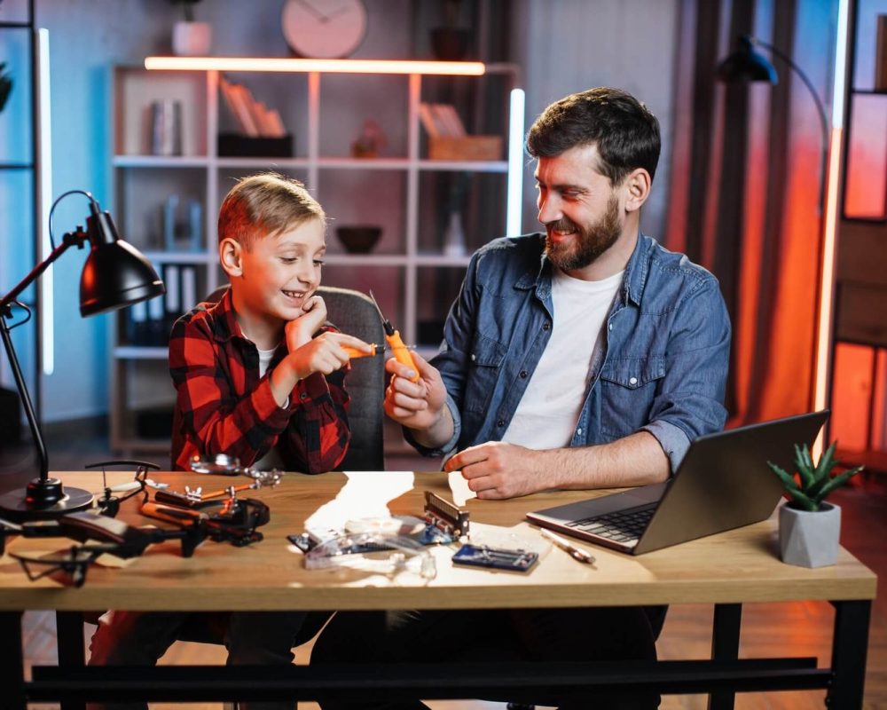 Father and son happily work on a drone project at a desk, surrounded by tools and a laptop. The scene conveys a sense of learning and bonding.