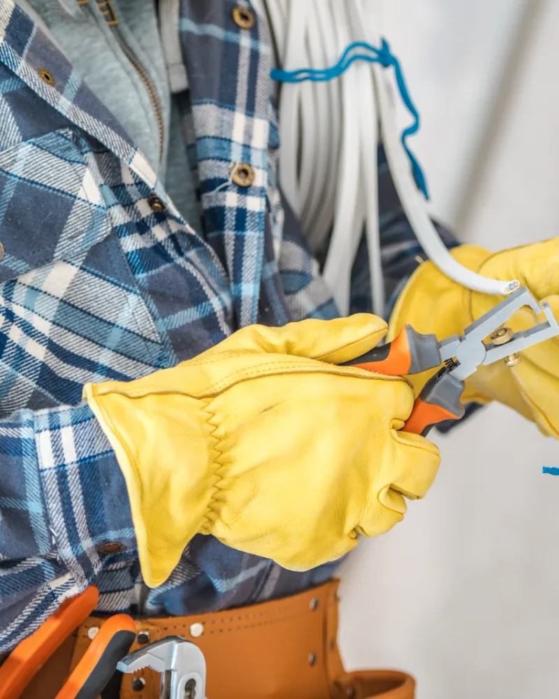 A person wearing yellow gloves and a blue plaid shirt uses pliers to work on electrical wiring inside a wall. A tool belt and cables are visible, conveying focus and precision.