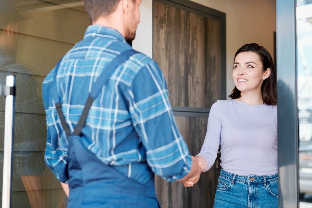 A woman in a light purple top and jeans smiles as she shakes hands with a man in a blue plaid shirt and overalls at a doorway, conveying friendliness.